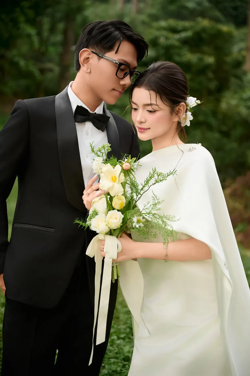 Cô dâu cầm bó hoa trắng vàng đứng cạnh chú rể đeo kính