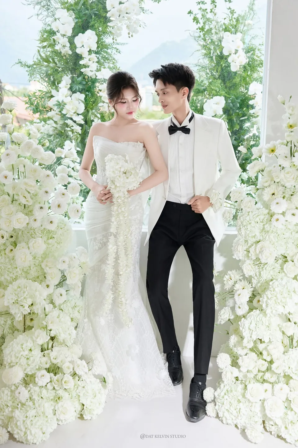 Cặp đôi đứng giữa hai mảng hoa trắng trước cửa kính