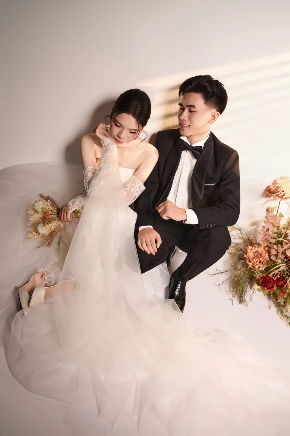 Cặp đôi ngồi trên nền trắng có bóng nắng, váy cưới trải rộng