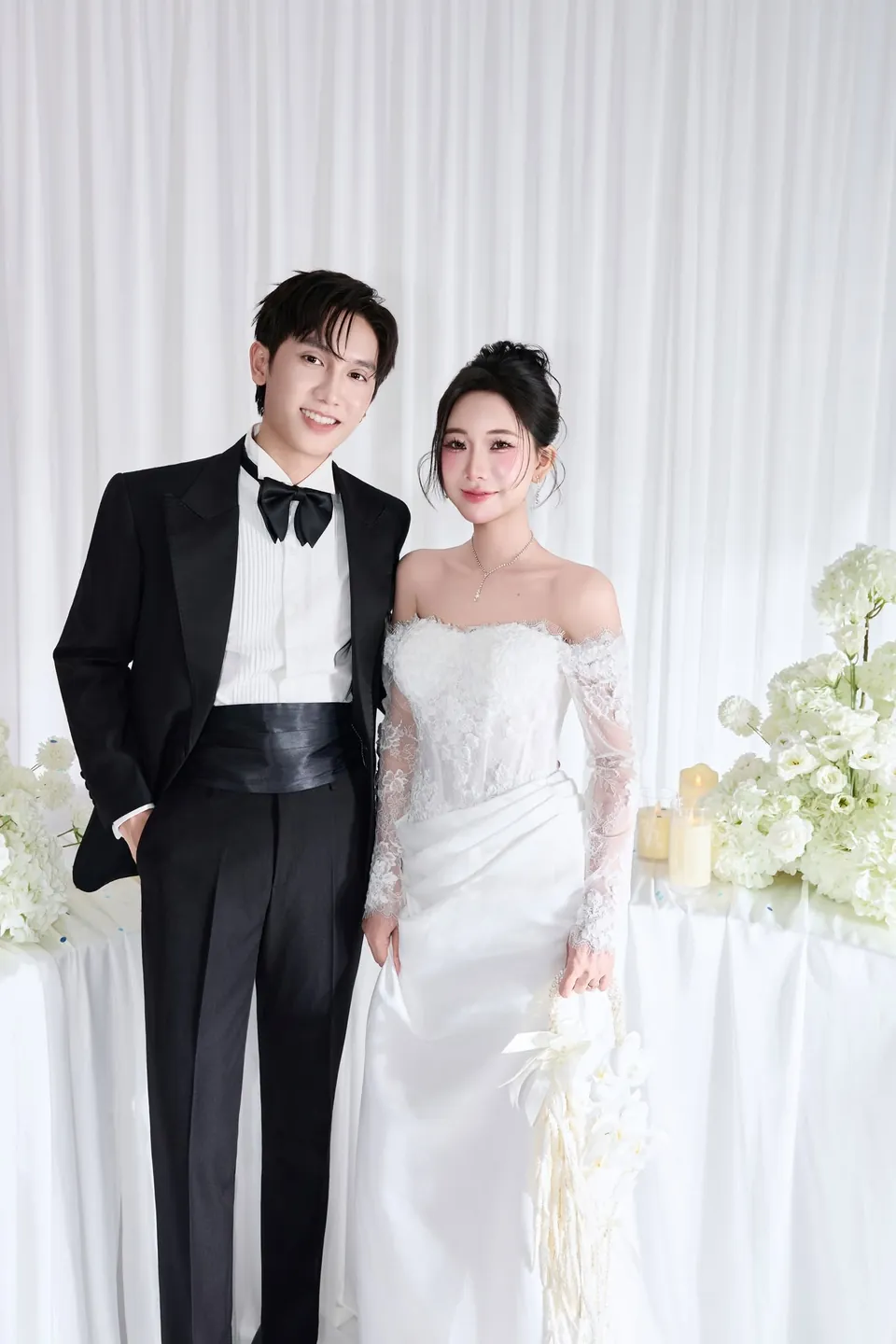 Cô dâu váy ren và chú rể tuxedo đen đứng cạnh nhau mỉm cười