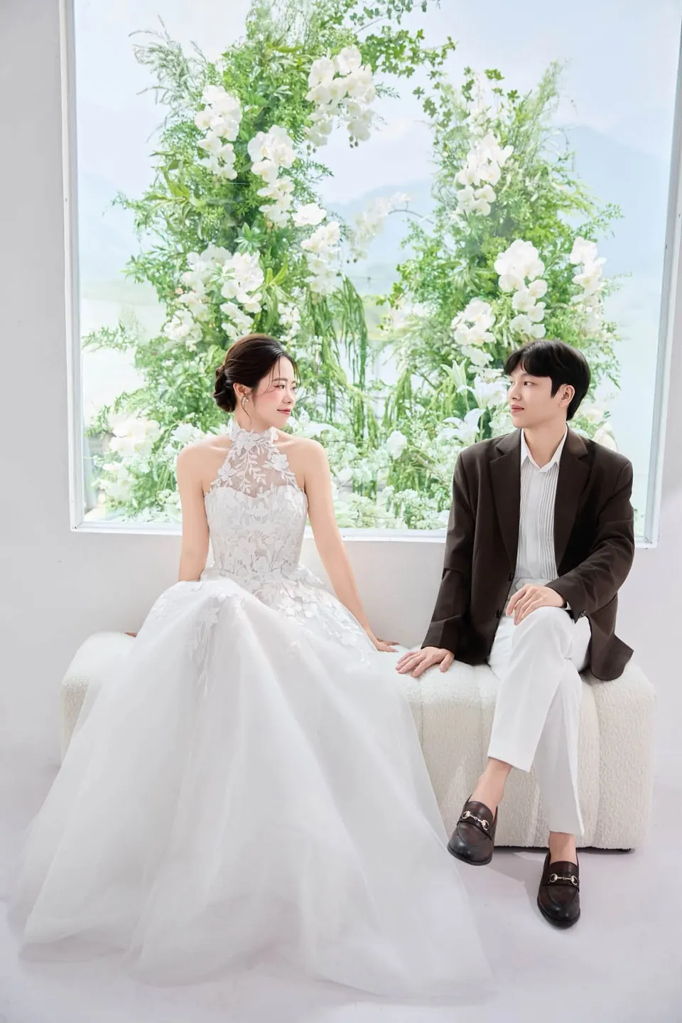 Cô dâu và chú rể ngồi trên sofa trắng trước khung cửa sổ đầy hoa