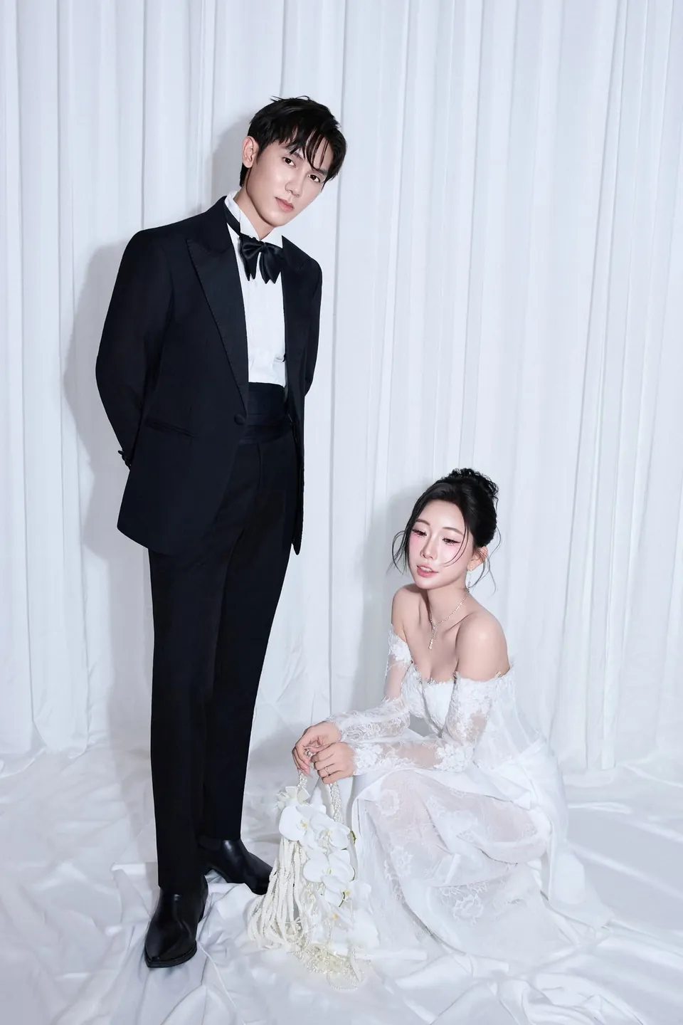Chú rể tuxedo đen đứng, cô dâu váy ren trắng ngồi trên nền vải trắng