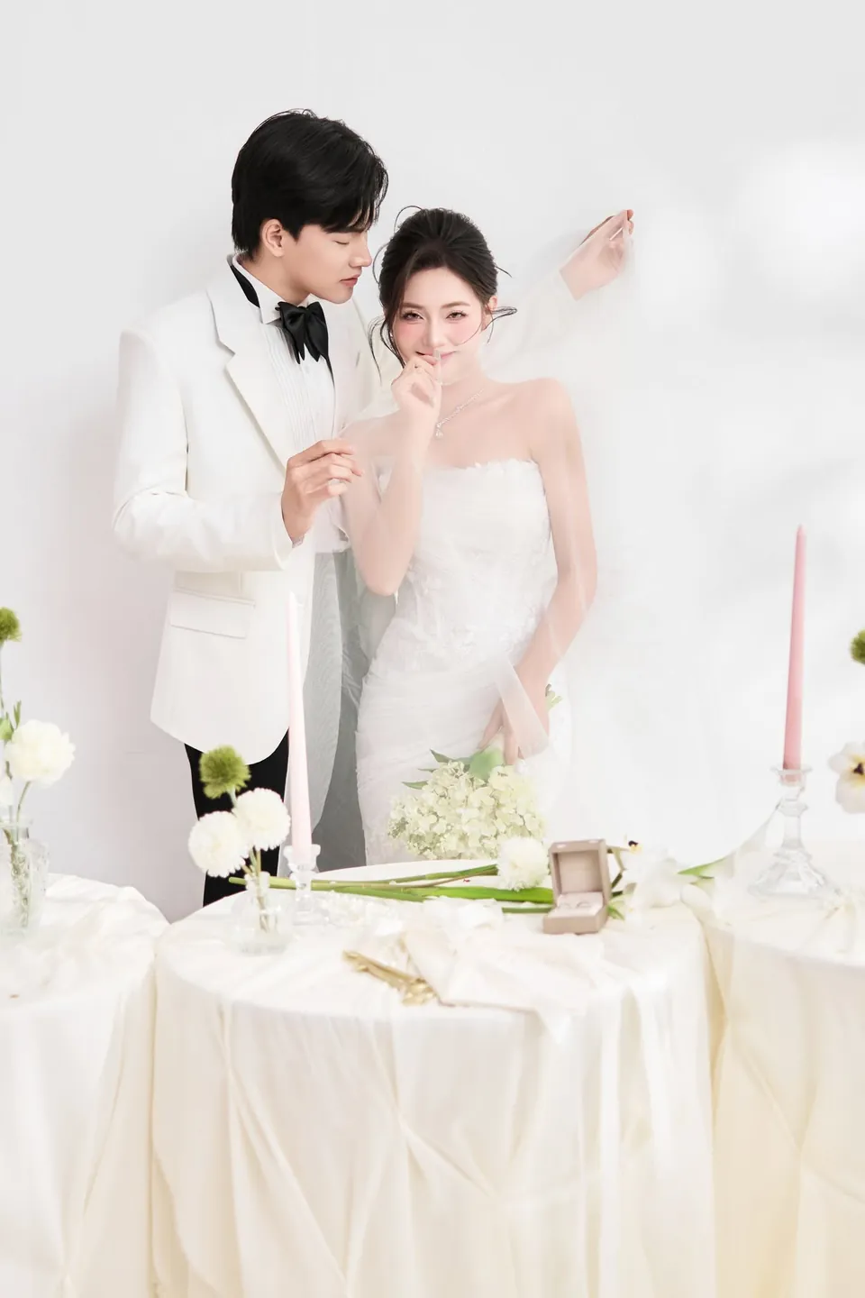 Cô dâu che miệng cười, chú rể nhìn nghiêng bên bàn tiệc trắng