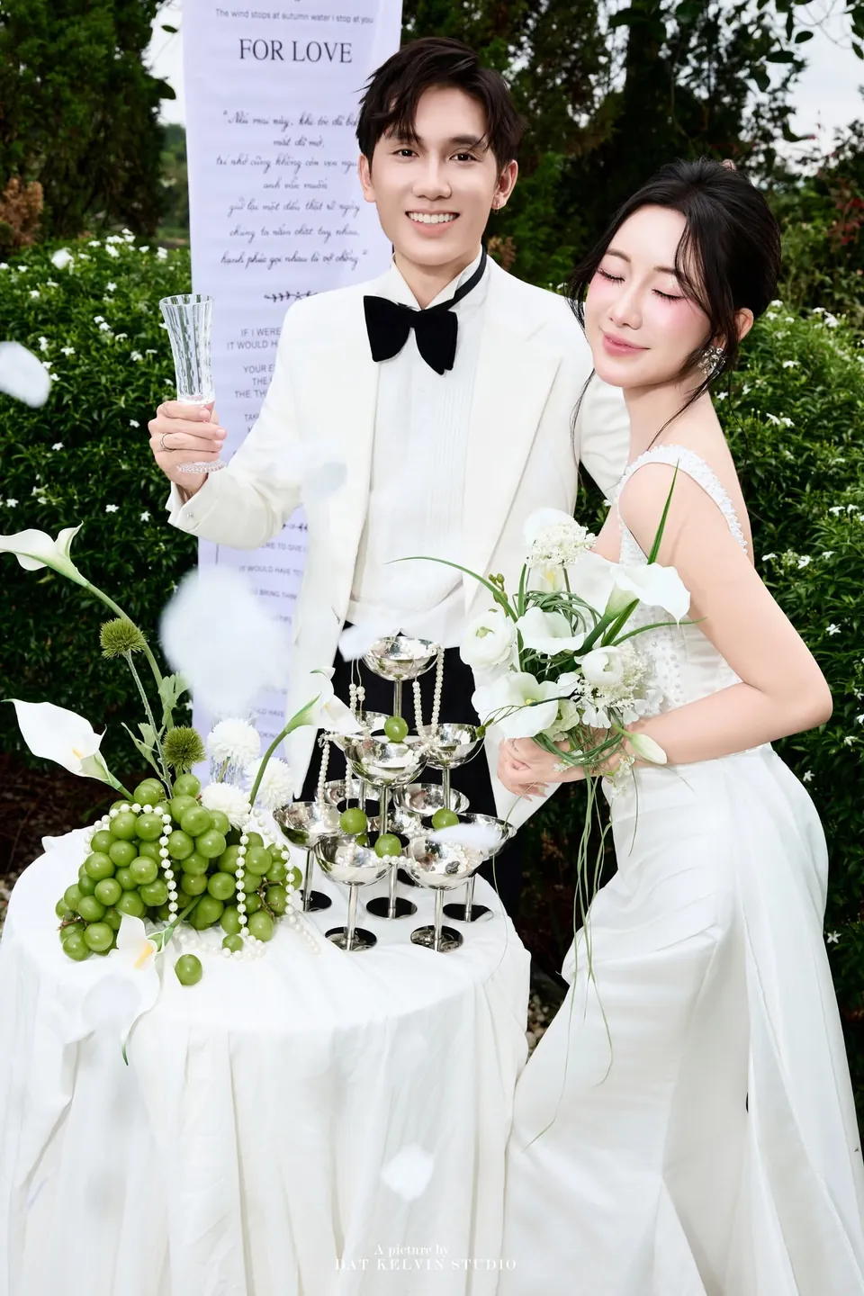Cô dâu tựa vào chú rể, phía sau là tấm băng lời thề For Love
