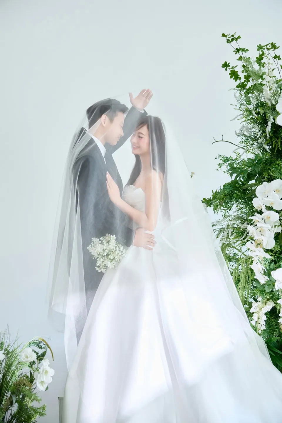 Cặp đôi đứng dưới tấm khăn voan dài bên mảng hoa trắng