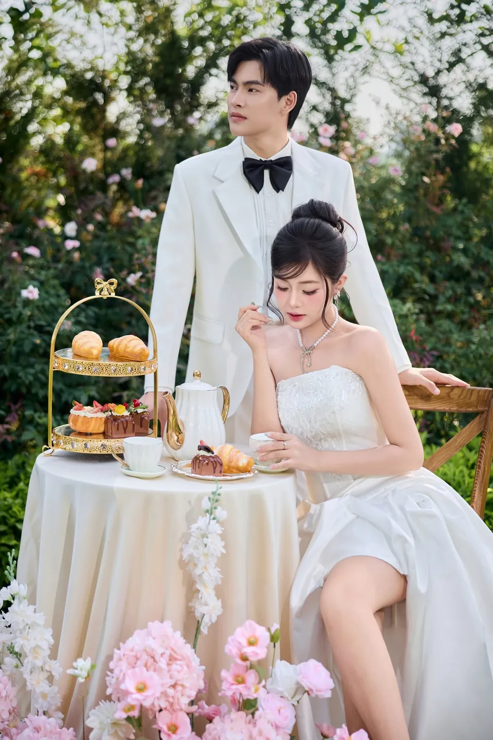 Chú rể đặt tay lên vai cô dâu đang ngồi cạnh bàn bánh ngọt
