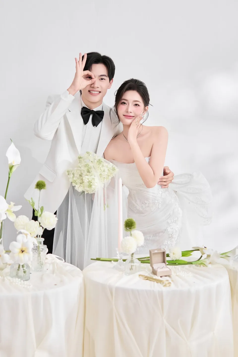 Chú rể vest trắng tạo dáng tinh nghịch, cô dâu cười bên bàn hoa trắng