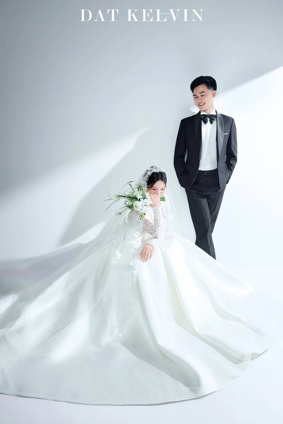 Cô dâu xoay tà váy dài trên nền trắng, chú rể đứng phía sau