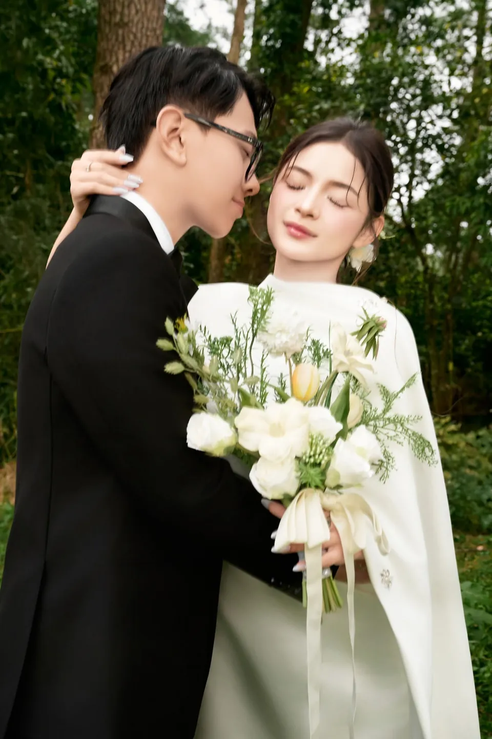 Chú rể vest đen ôm cô dâu váy satin trắng giữa rừng cây