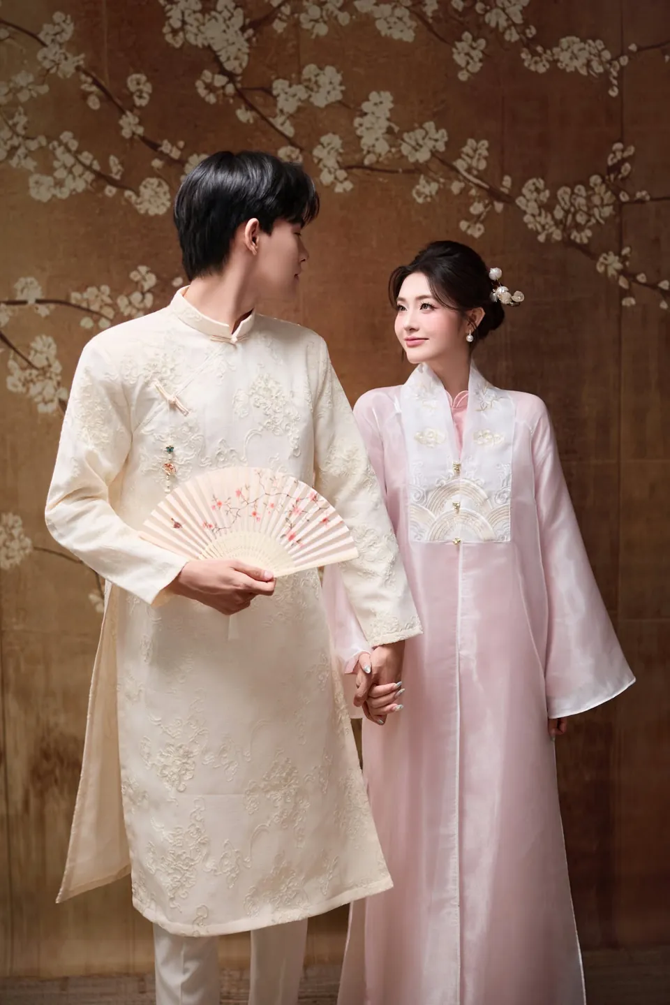 Cô dâu áo dài hồng và chú rể áo dài trắng ngà cầm quạt đứng trước phông tranh hoa