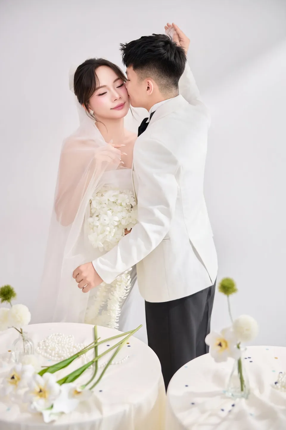 Chú rể ôm cô dâu từ phía sau giữa phòng chụp trang trí hoa trắng