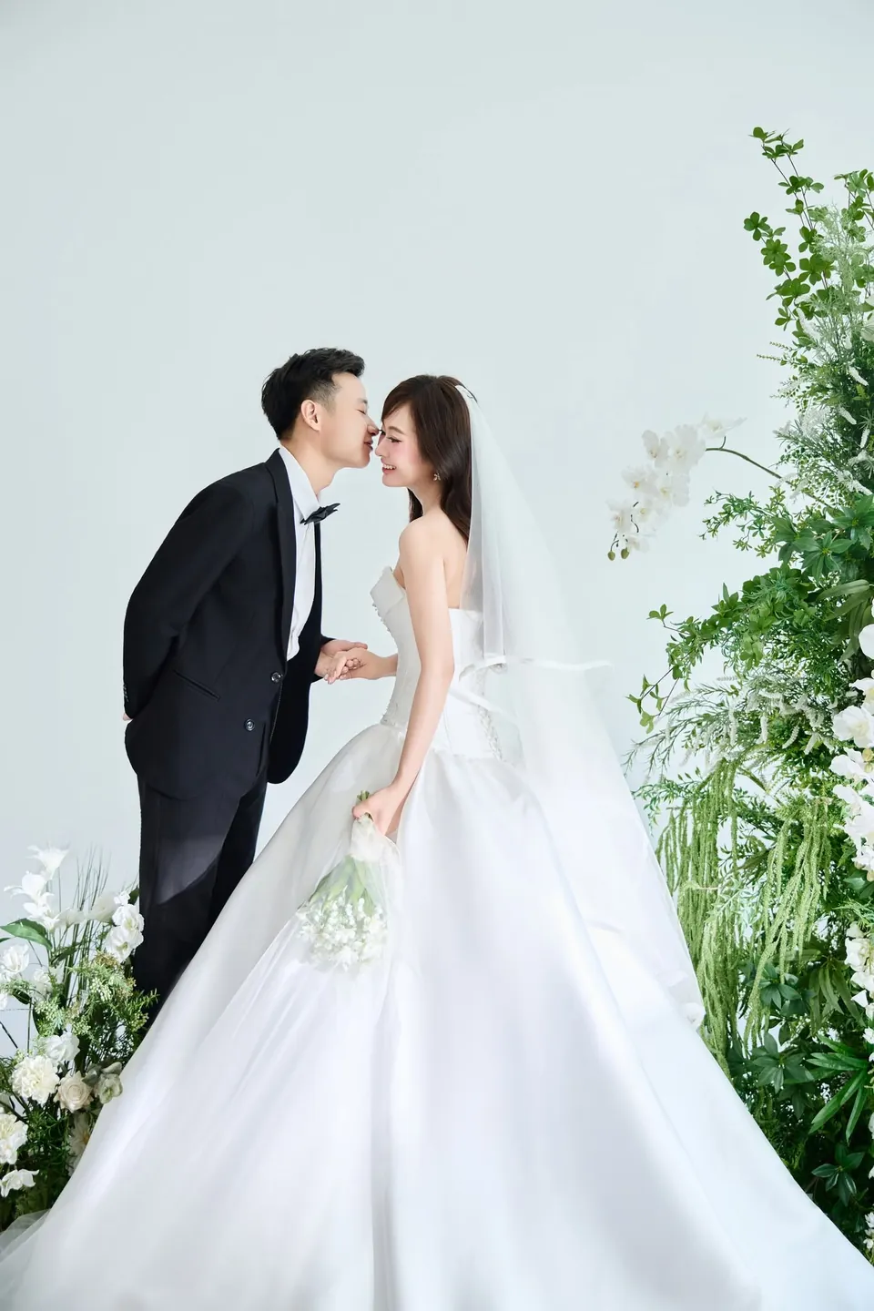 Chú rể vest đen hôn má cô dâu cầm bó hoa trắng