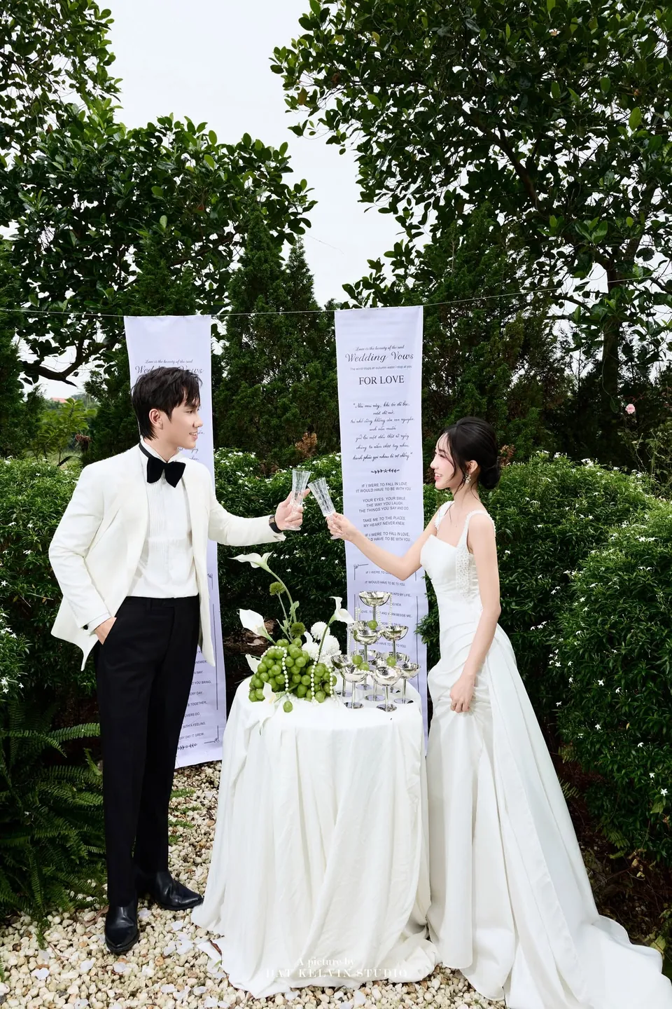 Cô dâu và chú rể đứng hai bên bàn tiệc trắng giữa khu vườn
