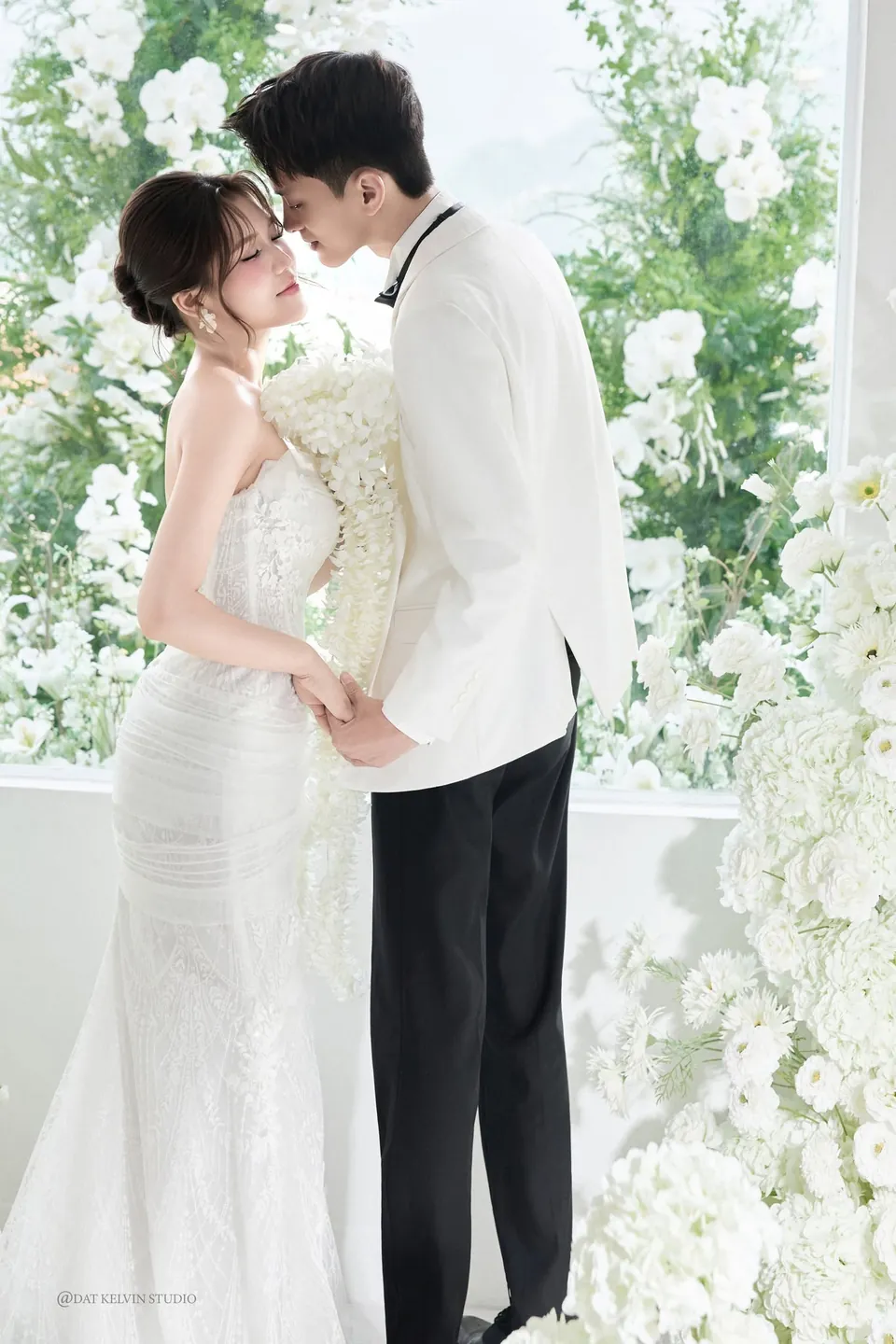 Chú rể ôm cô dâu bên bức tường hoa trắng