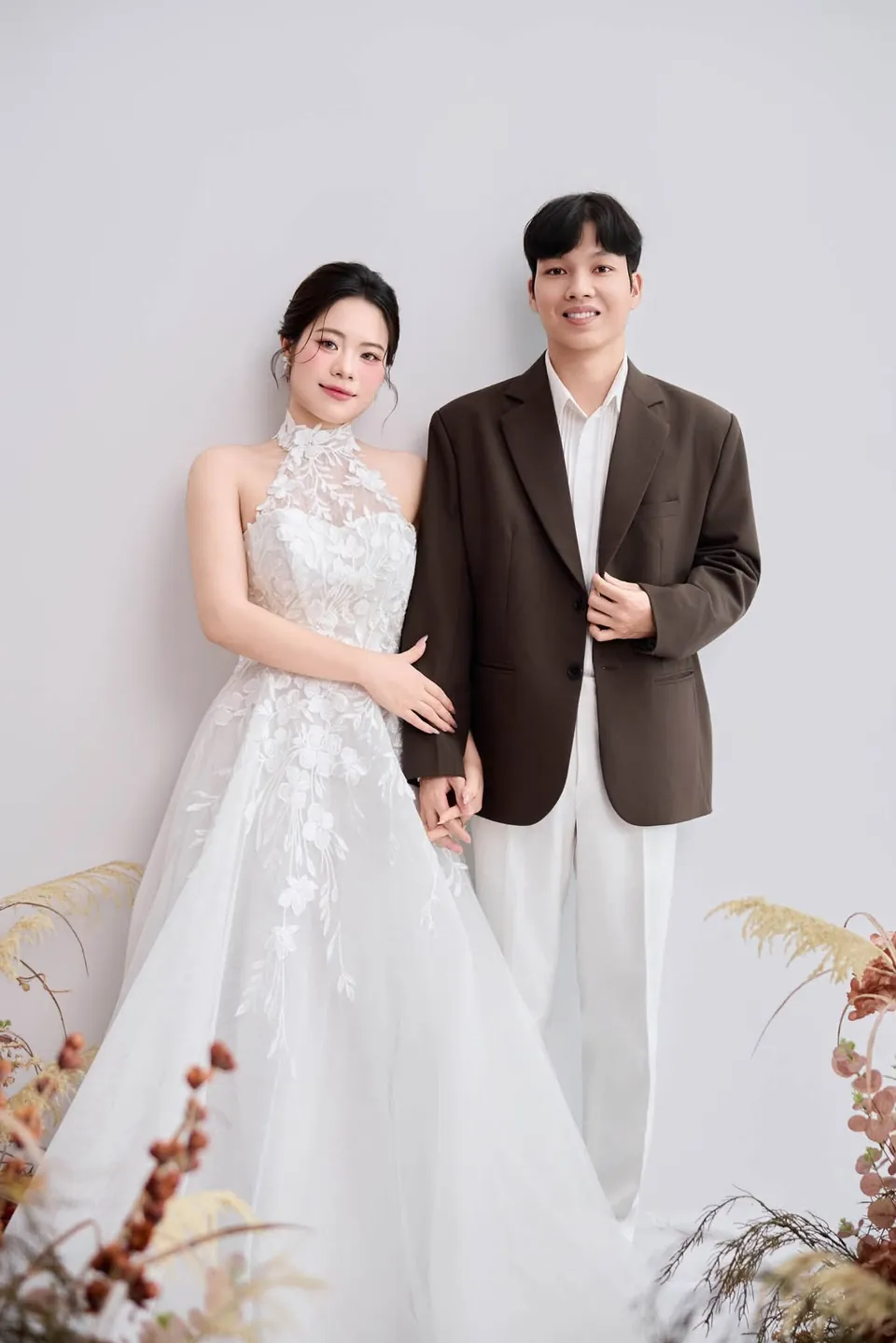 Cô dâu váy cổ yếm ren và chú rể vest nâu đứng trong phòng chụp nền trắng