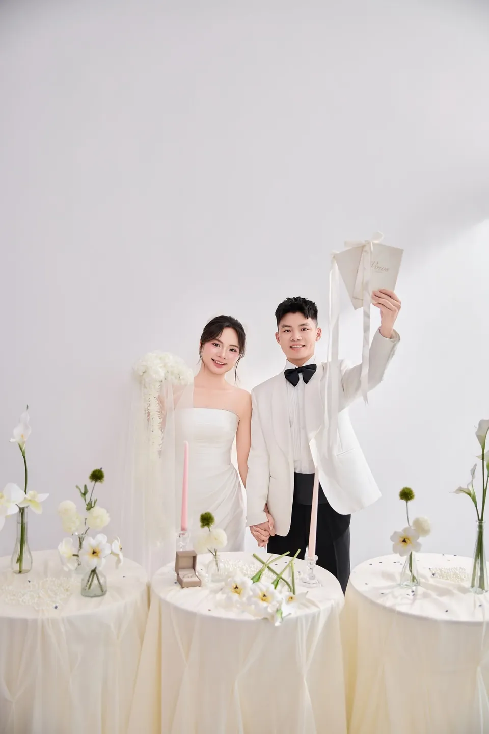 Cô dâu và chú rể đứng sau dãy bàn tròn bày hoa trắng