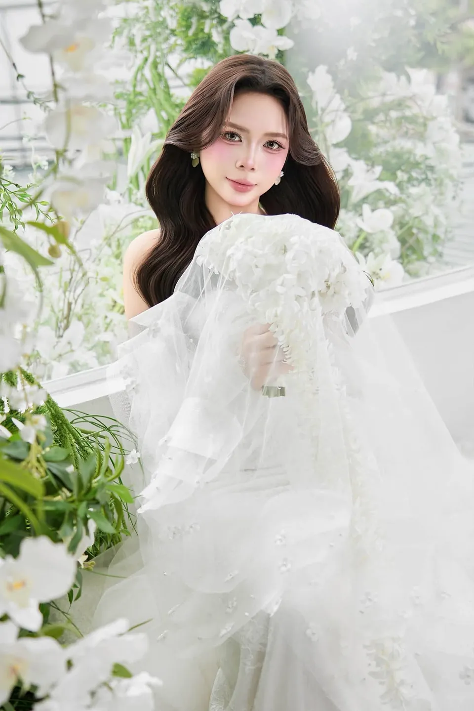 Cô dâu cầm khăn voan đính hoa trắng trước khung cửa sổ