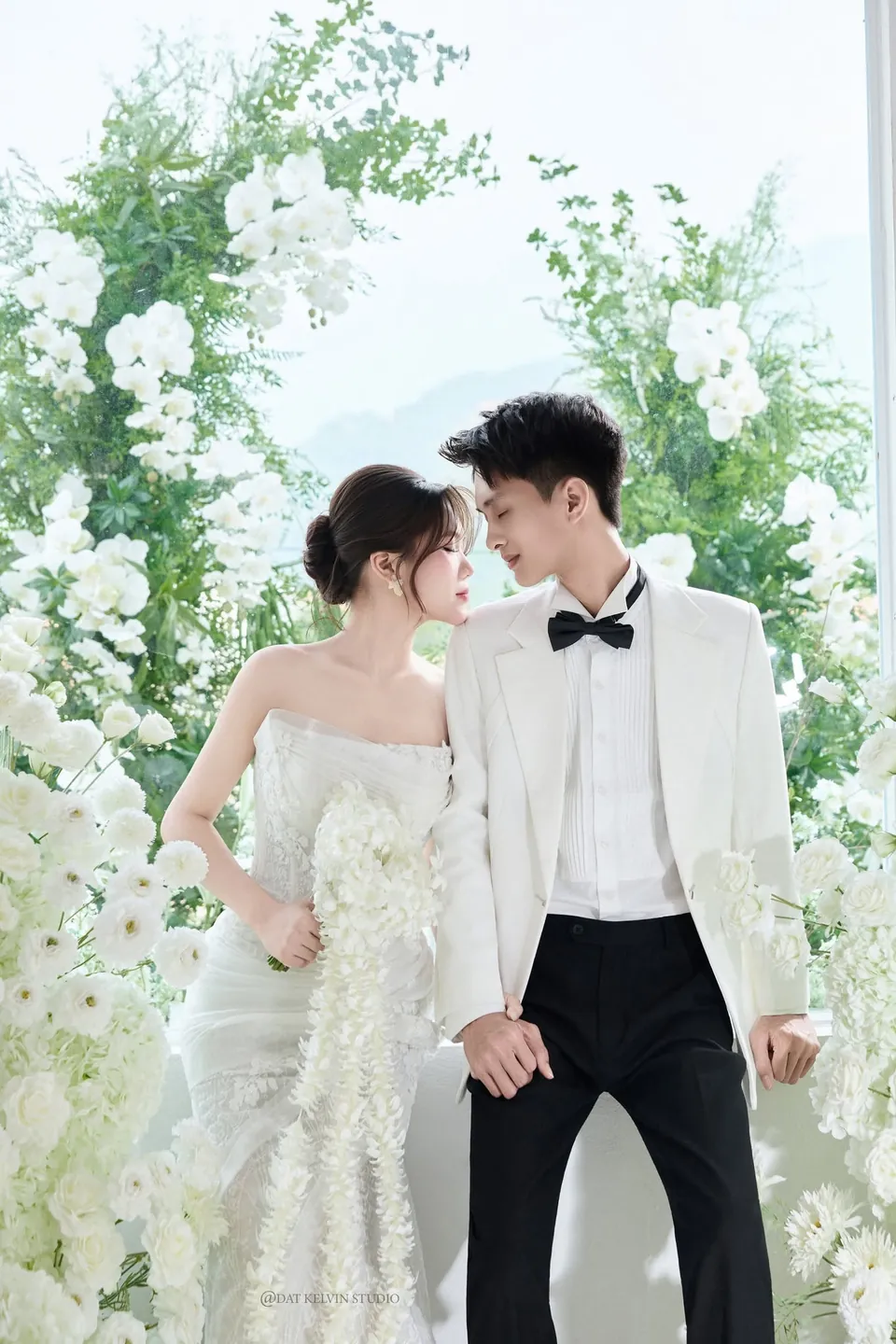 Cô dâu và chú rể vest trắng nhìn nhau giữa cổng hoa trắng