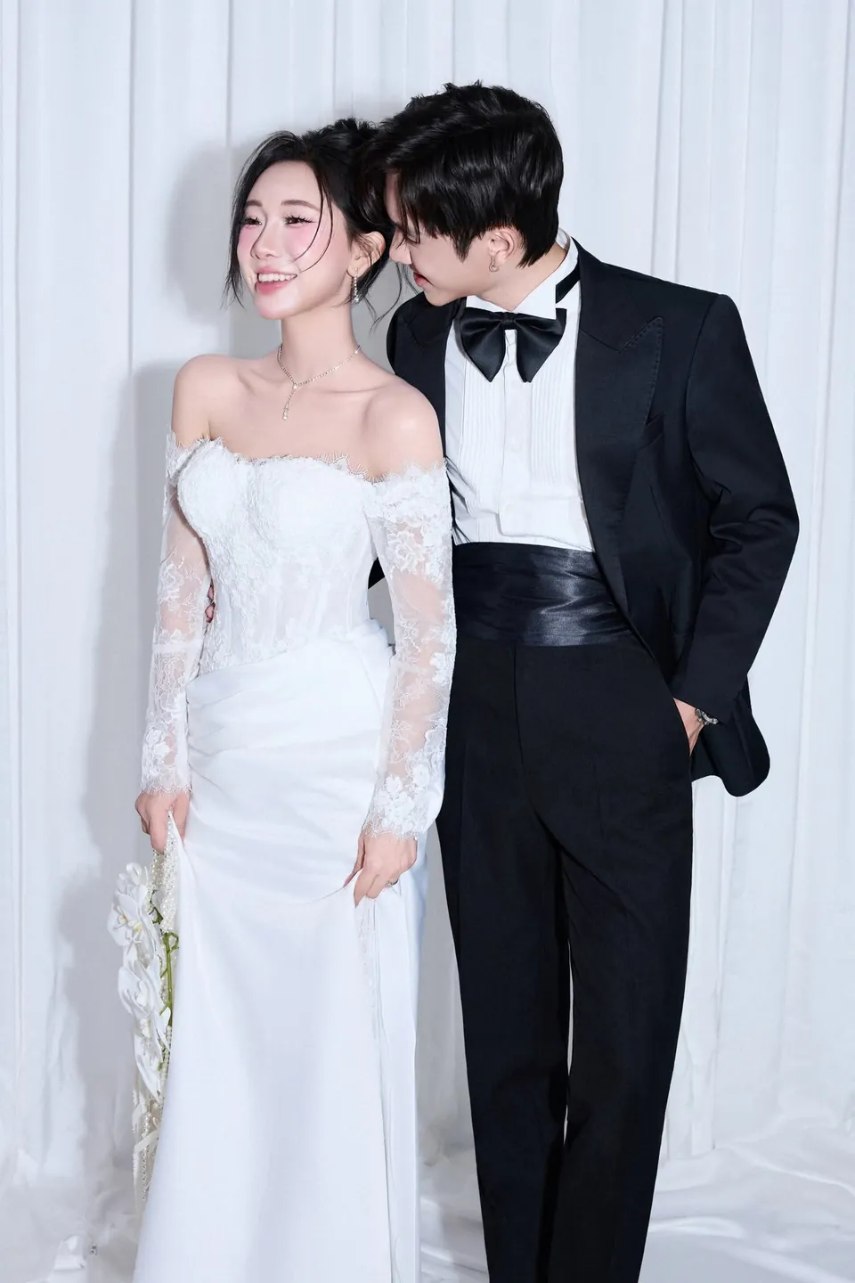Cô dâu cười tựa vào chú rể tuxedo đen trước rèm trắng