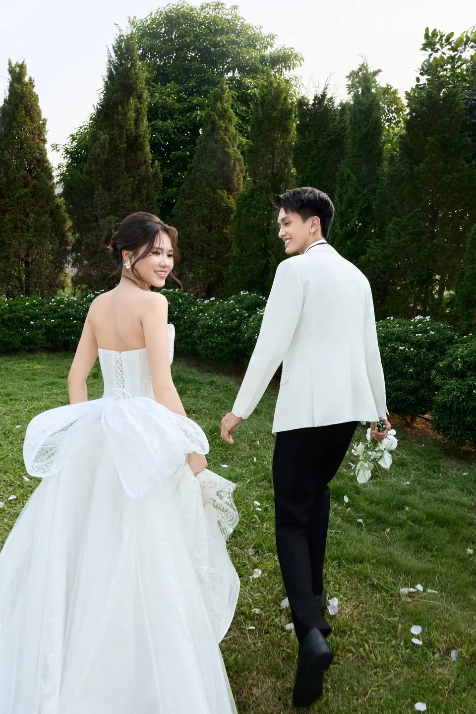 Cô dâu váy nơ lớn và chú rể vest trắng bước trên thảm cỏ
