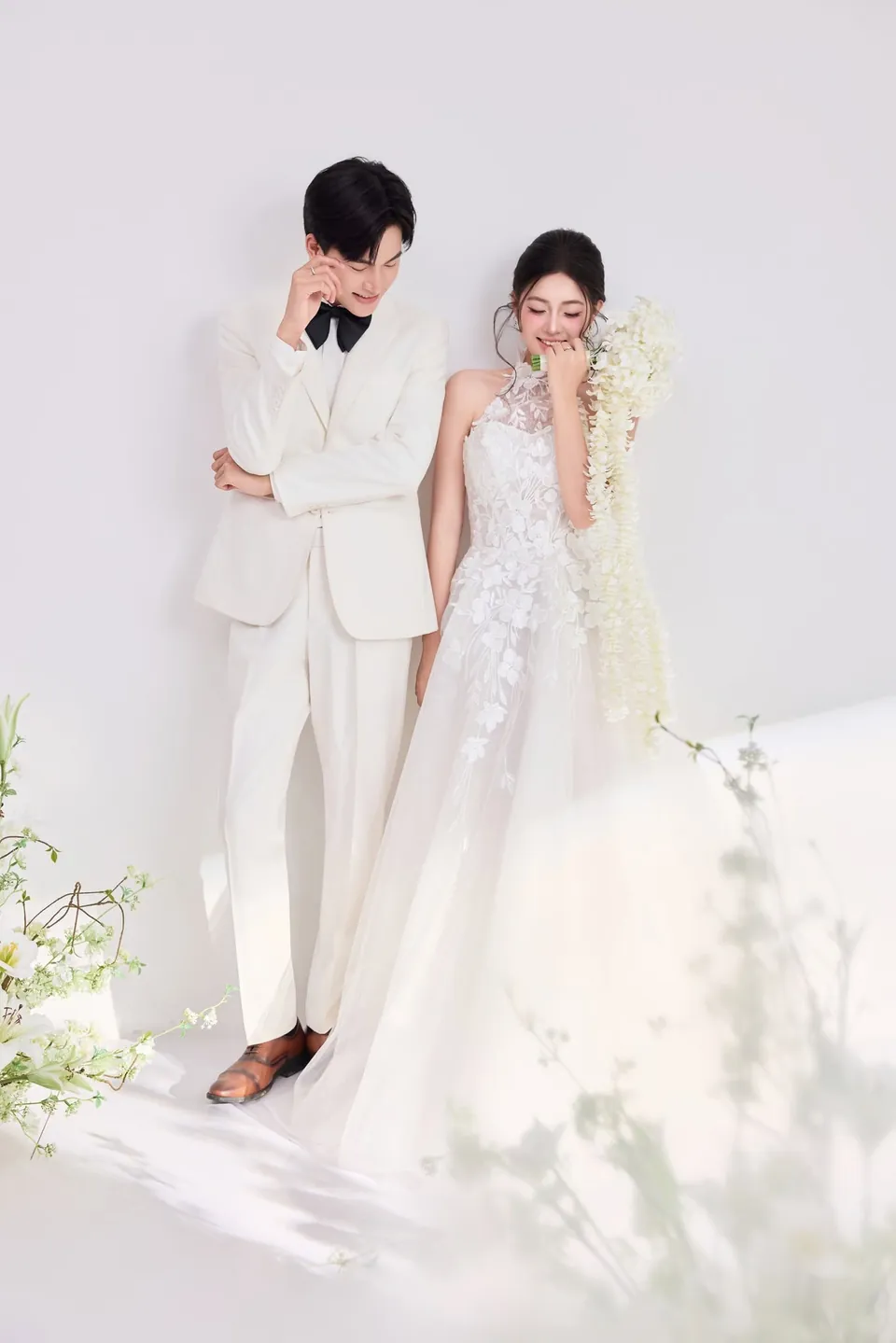 Chú rể vest trắng đứng cạnh cô dâu ôm bó hoa trắng