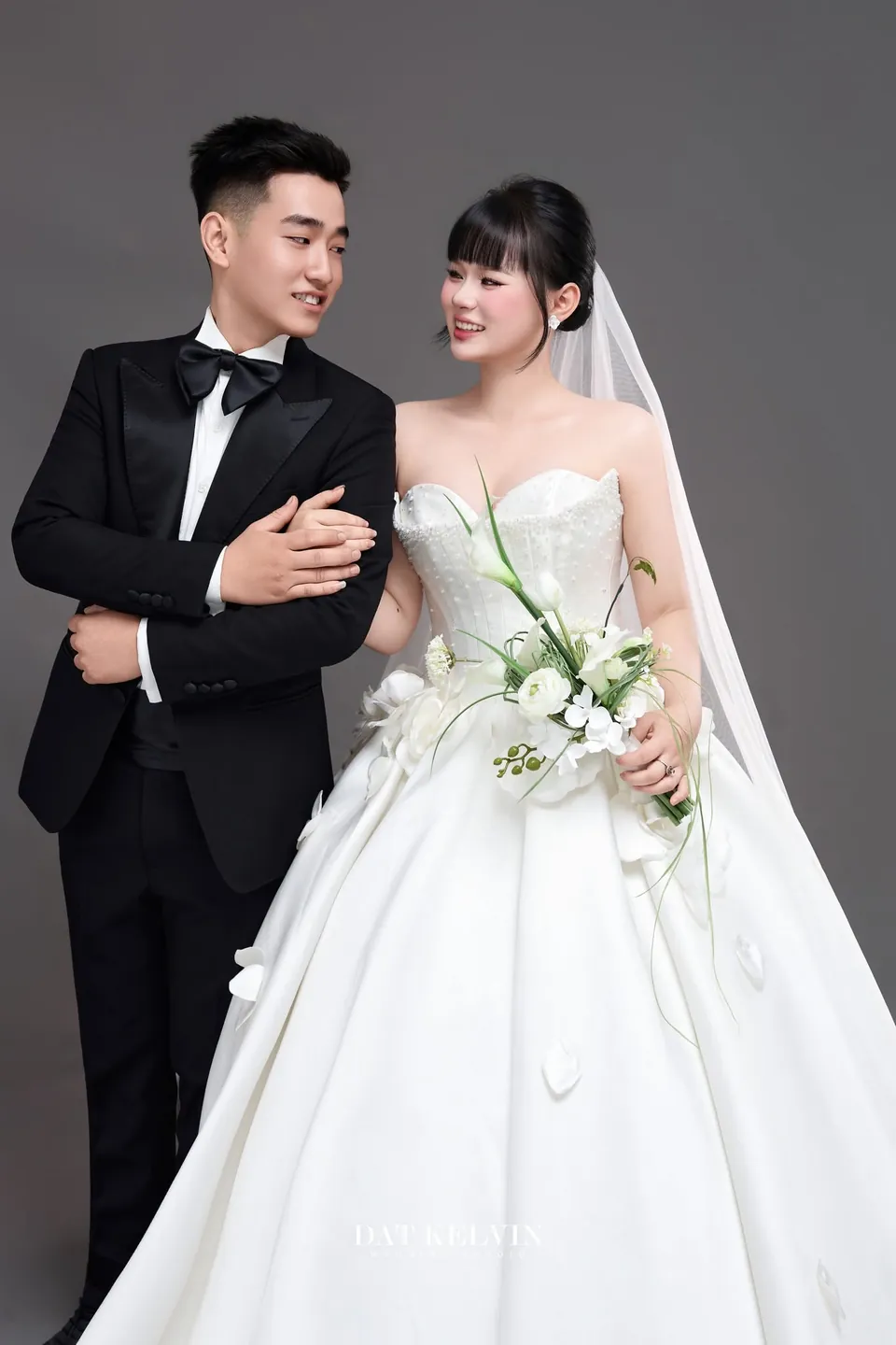 Chú rể vest đen ôm vai cô dâu váy công chúa trên nền xám