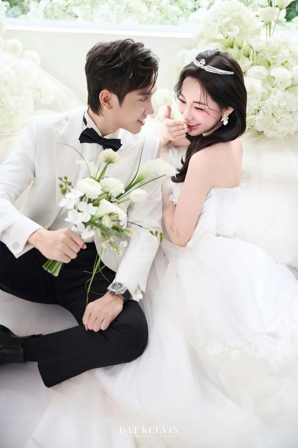 Chú rể vest trắng ôm hoa ngồi cạnh cô dâu đội vương miện
