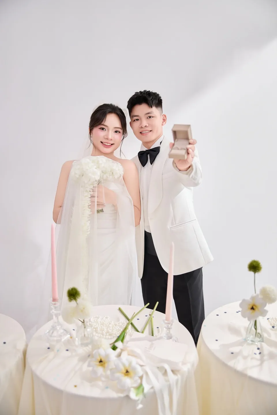 Cô dâu cầm bó hoa trắng, chú rể giơ hộp nhẫn cưới