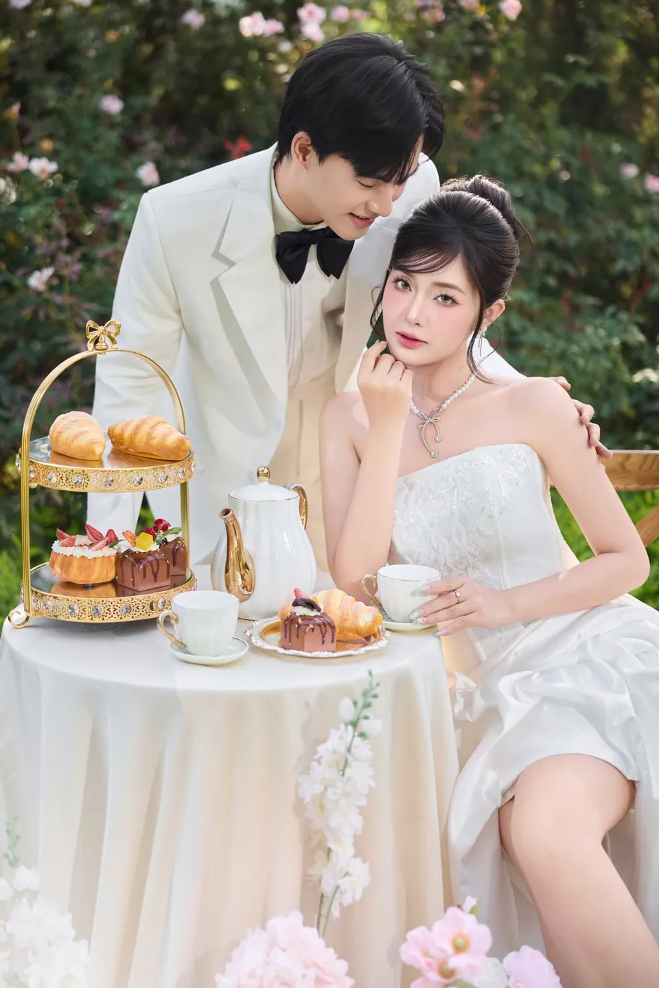 Cô dâu ngồi bên bàn tiệc trà có tháp bánh ngọt, chú rể vest trắng đứng phía sau