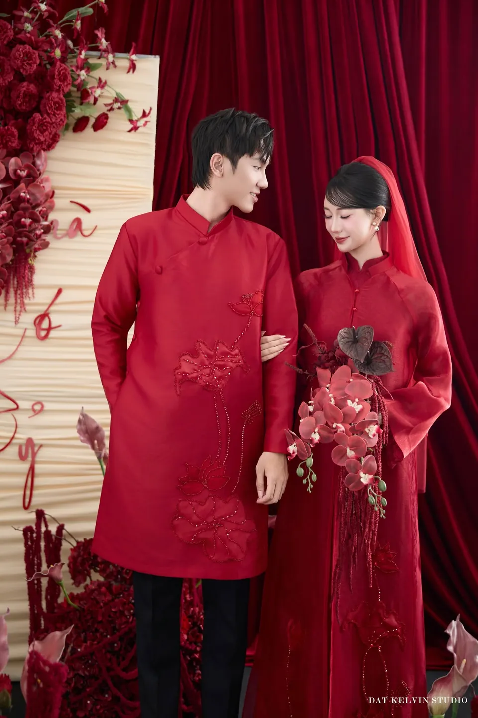Cặp đôi áo dài đỏ đứng cạnh nhau, cô dâu cầm bó hoa lan